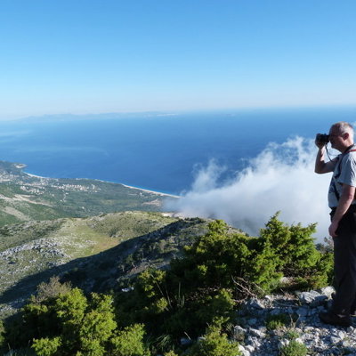Wandelaar met verrekijker op bergtop met uitzicht over de Albanese Rivièra en de Ionische Zee.