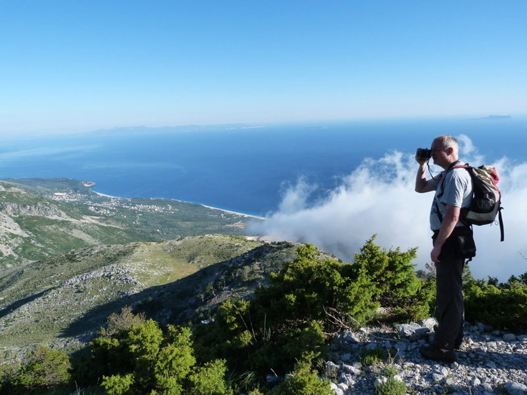Wandelaar met verrekijker op bergtop met uitzicht over de Albanese Rivièra en de Ionische Zee.