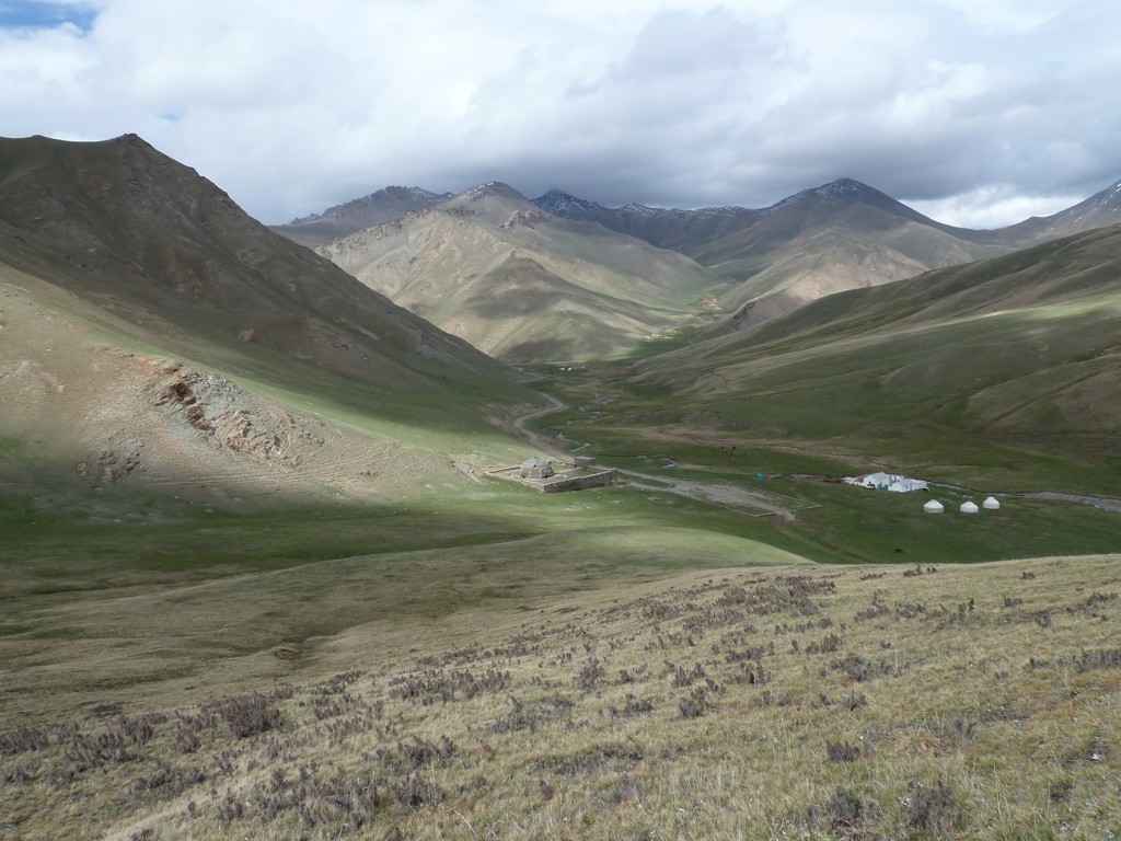 Groene bergvallei langs de Zijderoute met kronkelende rivier en enkele yurts onder bewolkte lucht.