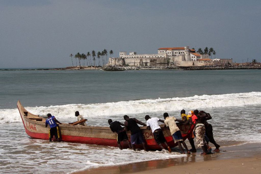 Vissers duwen een houten boot de zee in bij Cape Coast, met Fort Elmina op de achtergrond in Ghana.