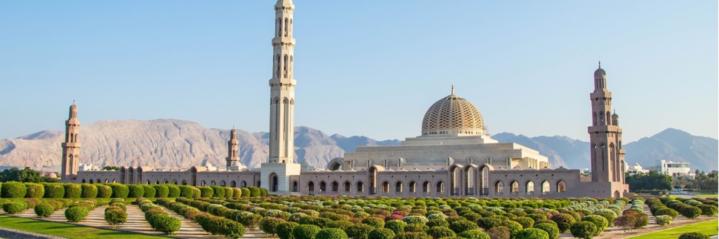 Sultan Qaboes-moskee in Muscat, Oman, met koepel, minaretten en tuinen tegen een bergachtergrond.