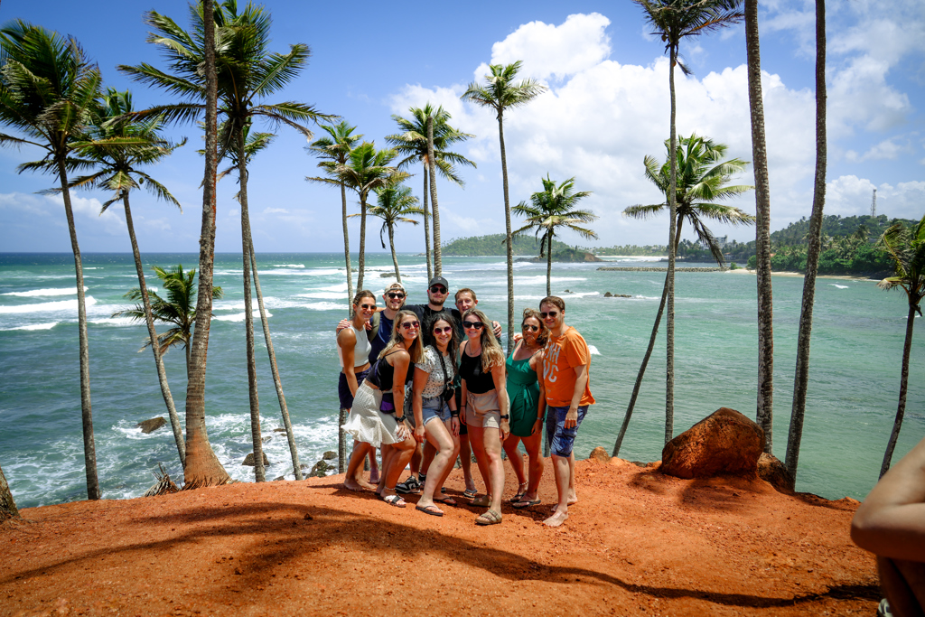 Groep 22-35ers poseert bij palmbomen aan de kust van Sri Lanka met uitzicht op zee en strand.