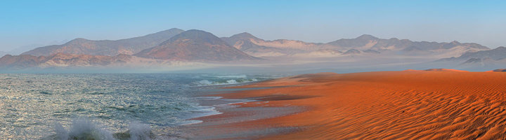 Rode zandduinen langs de Atlantische Oceaan bij de Skeleton Coast in Namibië, met bergen op de achtergrond.