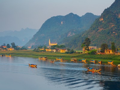 Rivierlandschap in Vietnam met kleurrijke bootjes, dorp en groene karstbergen op de achtergrond.