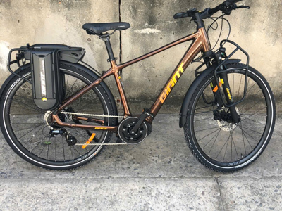 Bruine Giant e-bike met bagagedrager en accu, geparkeerd tegen een betonnen muur.
