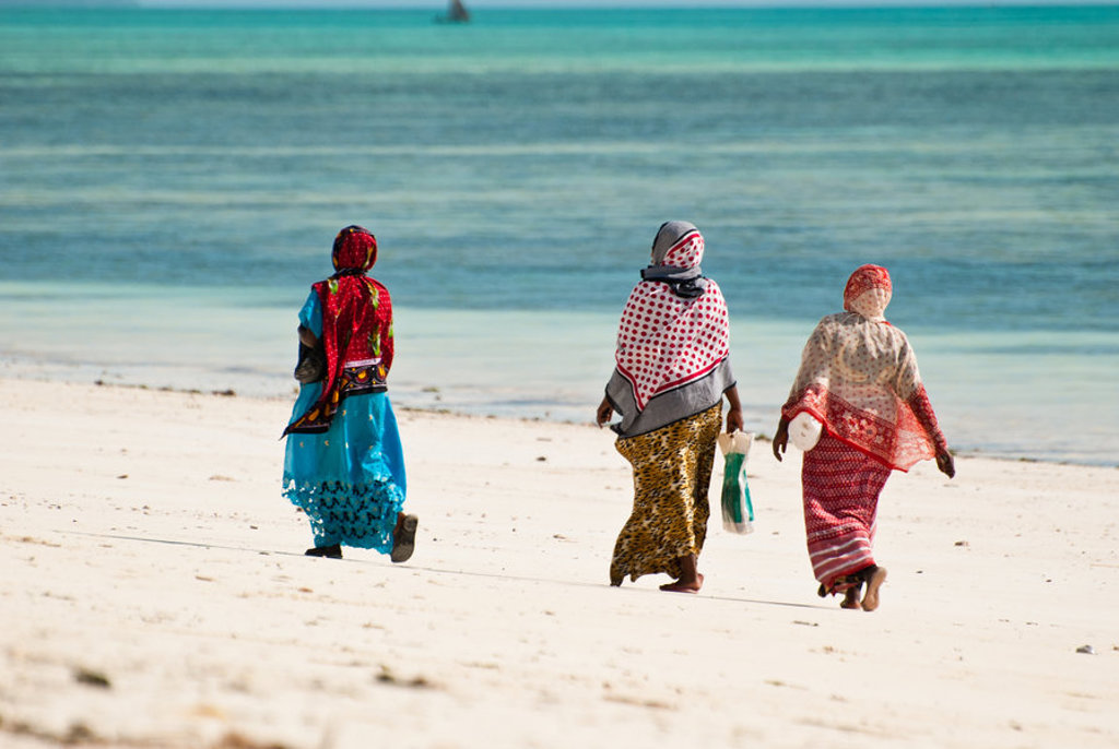 Drie vrouwen in kleurrijke kledij lopen langs het witte zandstrand van Zanzibar, met de blauwe oceaan op de achtergrond.