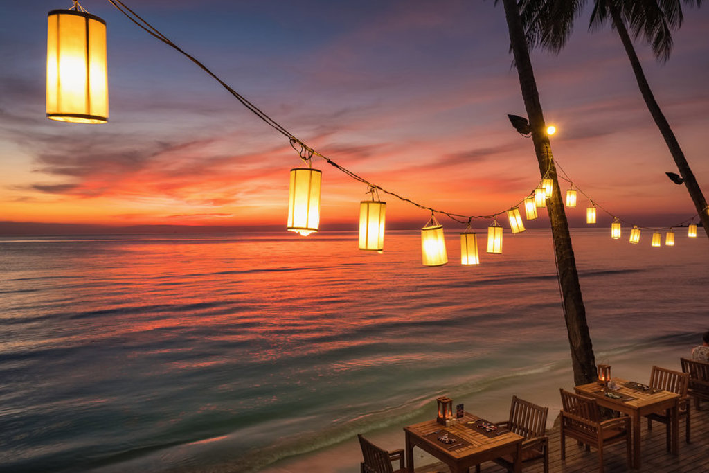 Sfeervol diner aan het strand bij zonsondergang in Thailand, met hangende lantaarns en zeezicht.