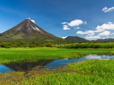 Arenal-vulkaan in Costa Rica met groene vlaktes en waterpartij onder een blauwe lucht.