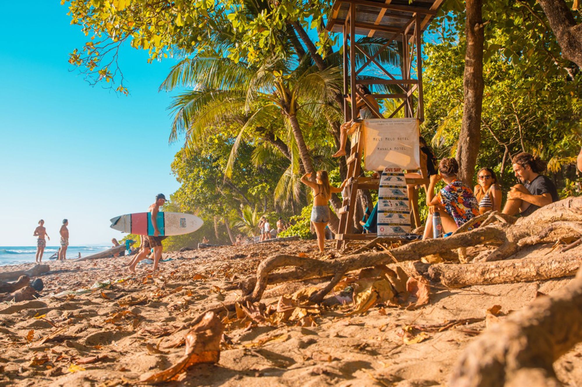 Surfers en strandgangers bij een strand met palmbomen en een lifeguardtoren in Costa Rica.
