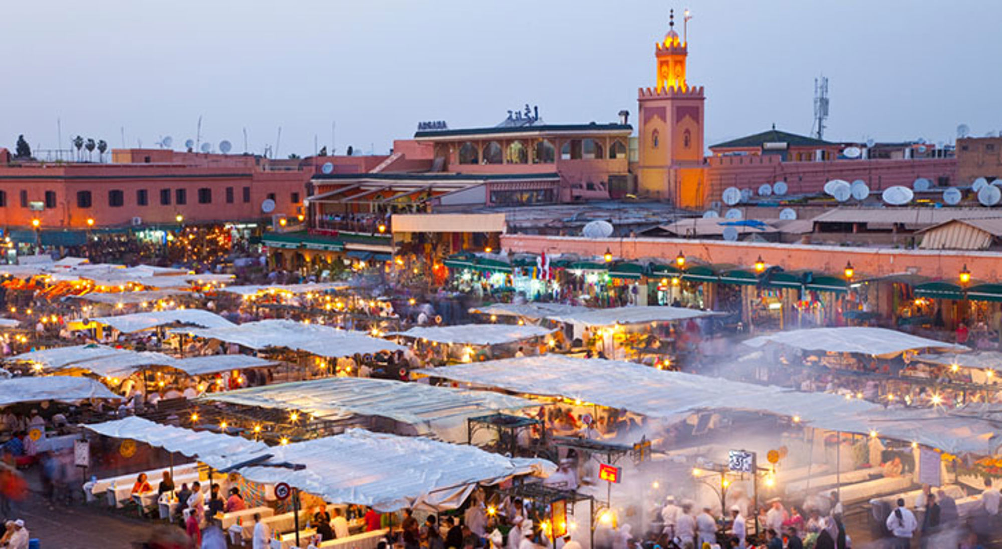 Jemaa el-Fna-plein in Marrakech bij schemering, met drukke markt en verlichte minaret op de achtergrond.