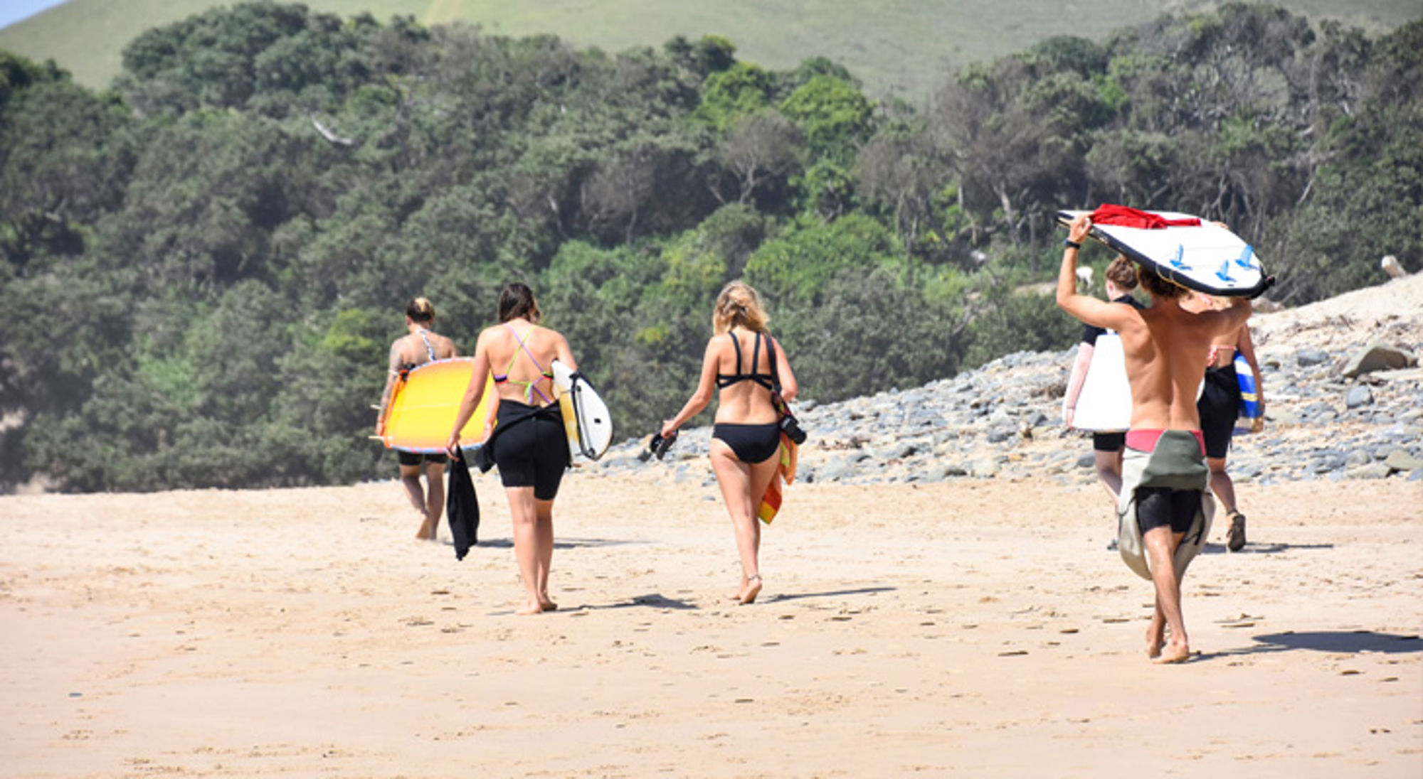 Surfen bij Coffee Bay Surfers lopen met surfboards over een zandstrand in Zuid-Afrika, met duinen en groen op de achtergrond.