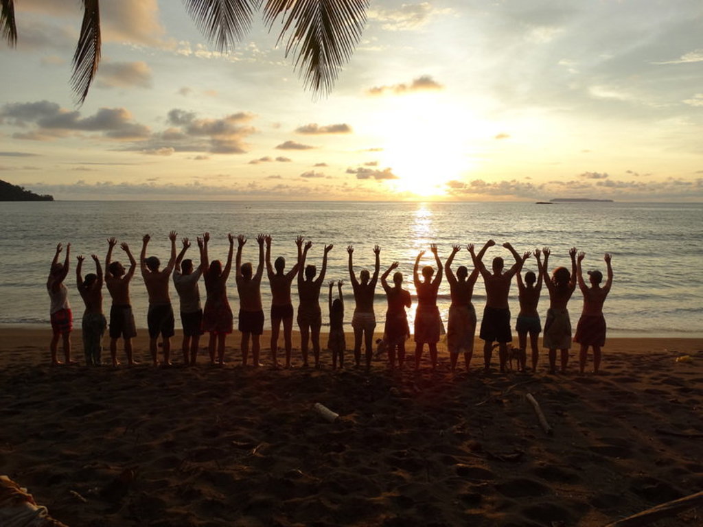 Groep reizigers op het strand in Costa Rica bij zonsondergang, met armen omhoog.
