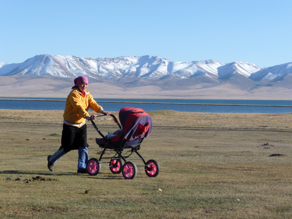 Vrouw duwt kinderwagen langs een bergmeer met besneeuwde bergen in Kirgistan tijdens groepsrondreis.