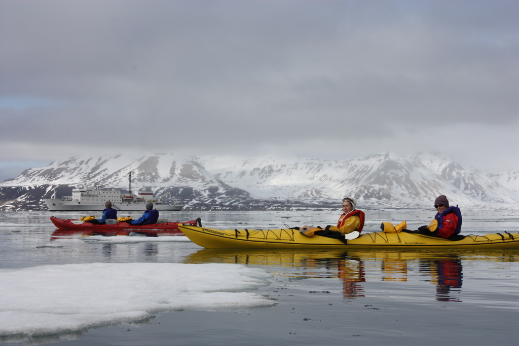 Kajakken tussen drijfijs bij Spitsbergen, met besneeuwde bergen en een expeditiecruiseschip op de achtergrond.
