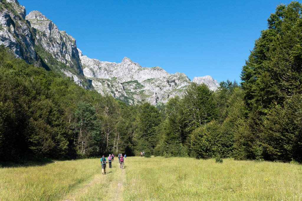 Wandelaars op een pad door een bergweide in de Peaks of the Balkan, met ruige bergtoppen op de achtergrond.