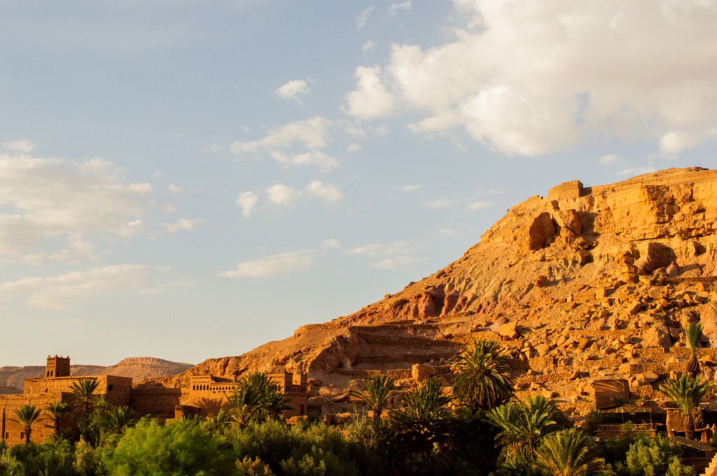 Kasbah bij Aït Ben Haddou in Marokko, met palmbomen en rotsachtige heuvel in avondlicht.